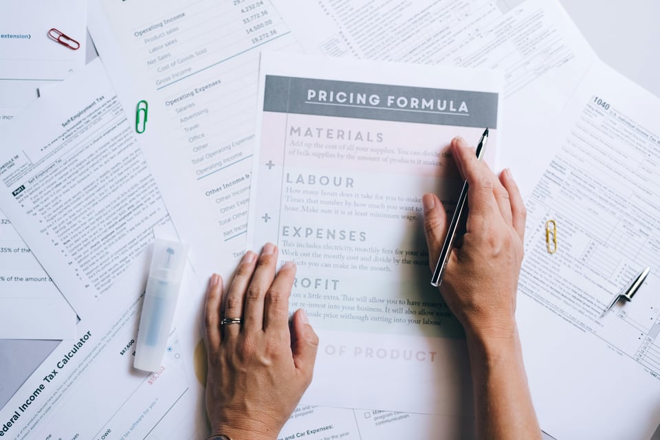 Hand with a pen over a pricing formula worksheet listing materials, labour, expenses, and profit