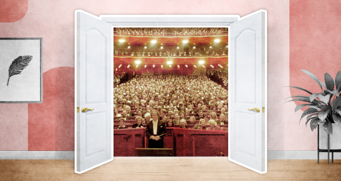 Double doors open to reveal a large auditorium filled with people seated, facing the stage, in a warmly lit theater.