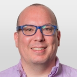 A person with blue glasses and a light purple shirt smiles against a plain background.