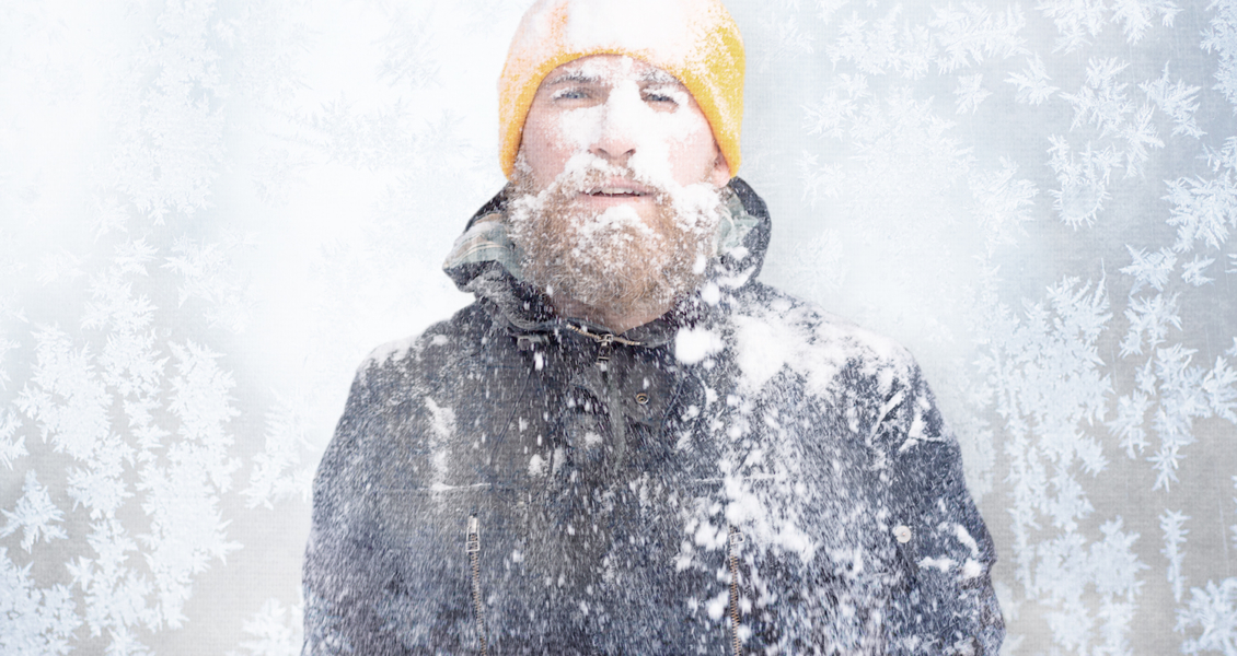 A person with a beard and yellow beanie stands in snowy weather, embodying the chill of a writer's block. Snowflakes cover their jacket and face, while frost patterns create an intricate backdrop.