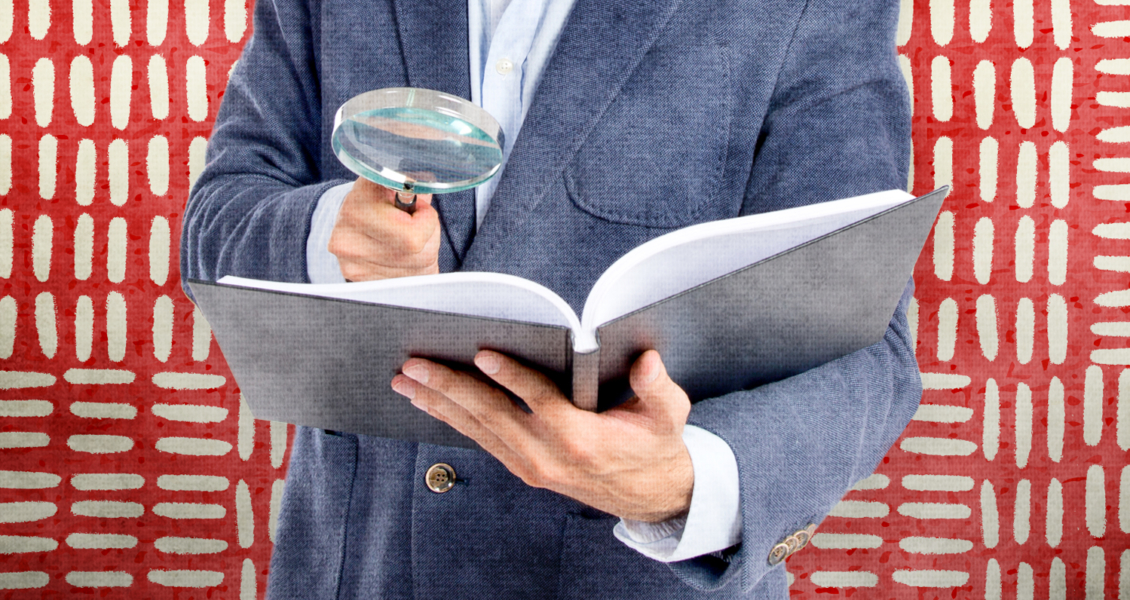 Person holding a magnifying glass and examining an open book in front of a red patterned background.