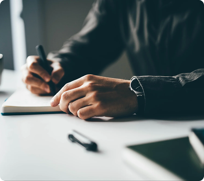 A person wearing a watch writes in a notebook at a desk. A pen is lying nearby.