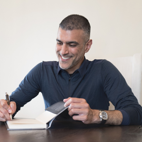 Man in dark blue shirt sitting at a table signing the inside of a book.