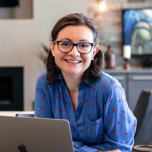 Brunette woman with glasses in blue top smiles while sitting with her laptop computer.