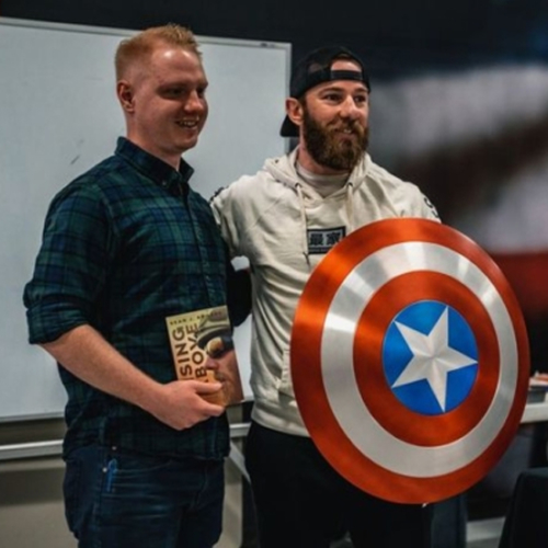 Two men standing beside each other--one holding a book, the other holding a Captain America sheild.